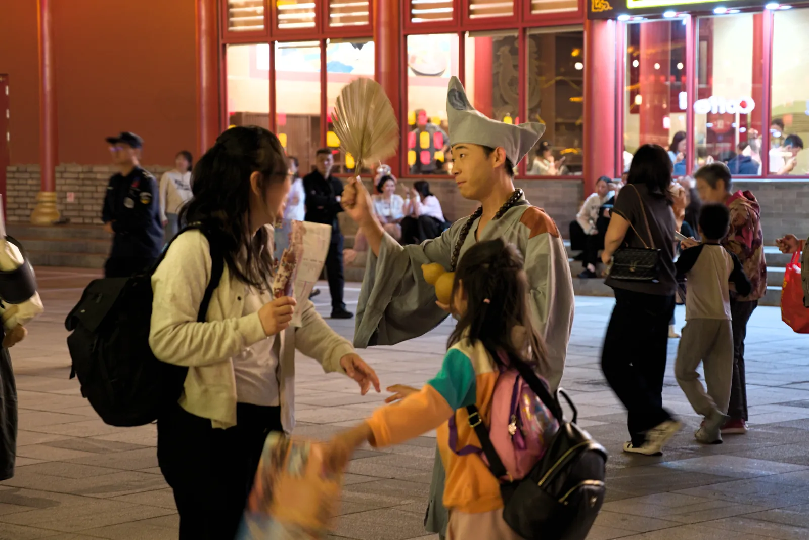 夜晚仿古街区的沉浸式互动现场：一身僧袍、手举蒲扇的济公扮相演员正与游客母女交谈，背景是灯光明亮的店铺和往来人群
