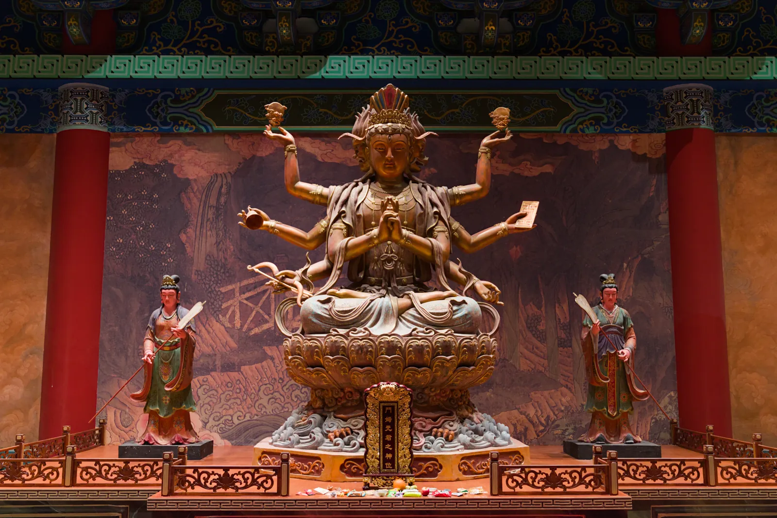 殿内供奉的多臂神像：头戴宝冠、结跏趺坐于莲花座上，两侧各立一名侍者，身后是山水壁画，殿内以红柱和青绿彩画为饰