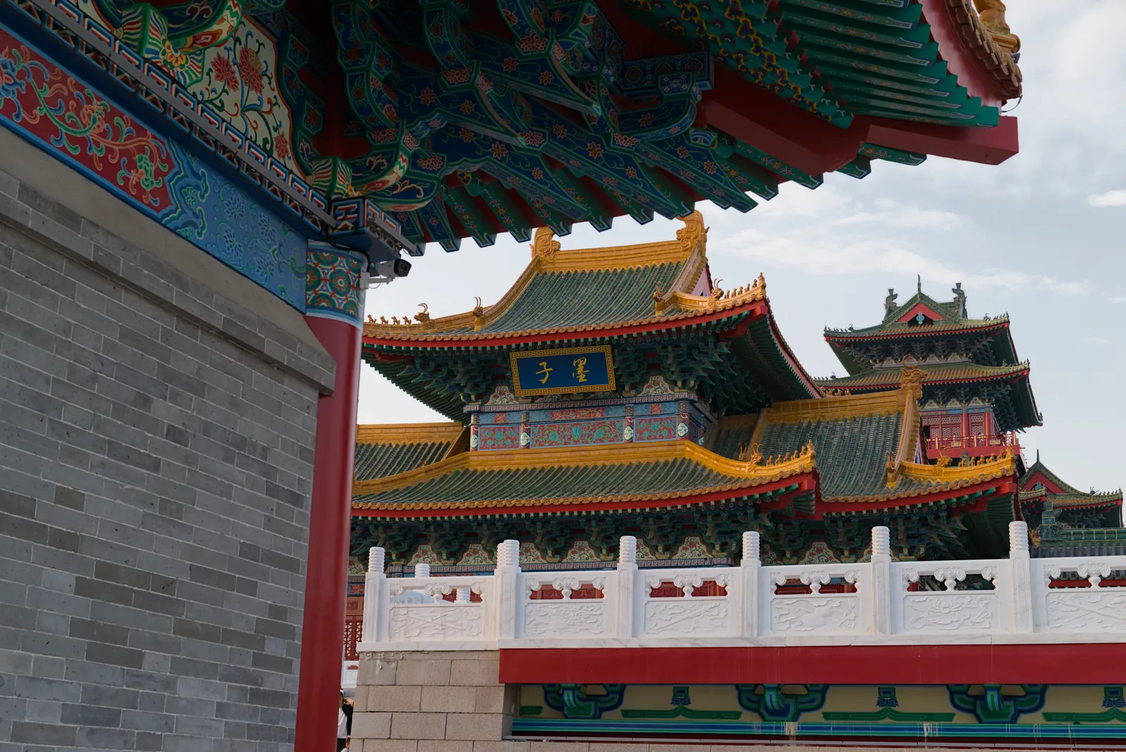檐下视角：一座重檐仿古建筑悬着蓝底金字的“墨子”匾额，屋面为绿琉璃瓦配黄色屋脊，前景是灰砖墙、红柱和汉白玉栏杆