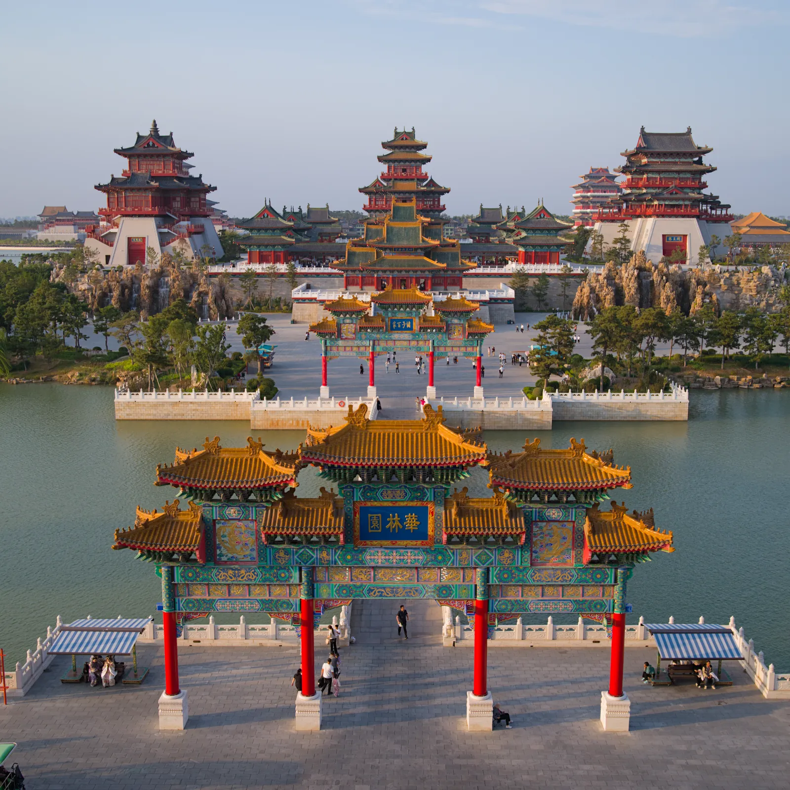 俯拍华林园风景区入口：正中是四柱三间的彩绘牌楼，蓝底金字匾额写有“华林园”，黄琉璃瓦顶配红柱，牌楼两侧是水面与石桥，远处是层层殿宇和假山。